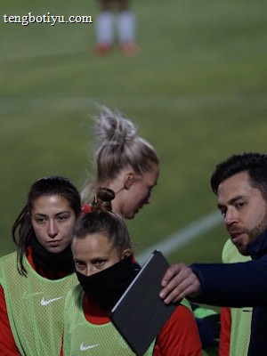 A group of individuals on a sports field are wearing green vests over red attire. One person is holding a tablet and seems to be discussing or showing something to the others. The atmosphere suggests a sports event, likely soccer, indicated by their clothing and the setting. The background is a grassy field.