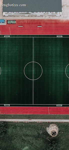 An aerial view of a large sports complex featuring a central green football field surrounded by a red running track. Adjacent to the track, there is a parking area, buildings, and pathways. Nearby, there are additional sports courts in different colors, including a blue swimming pool and orange basketball courts.