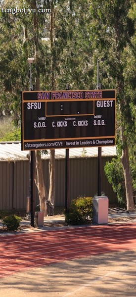 An outdoor scoreboard for San Francisco State is seen among trees and greenery. The board displays sections for shots on goal and corner kicks, with areas for both home and guest teams. It is situated near a running track with a building in the background.