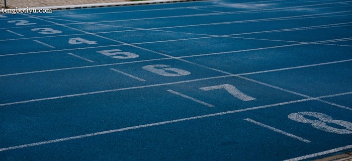 A sports track with multiple blue lanes, each marked with white numbers from 2 to 8, is visible. Adjacent to the track is a paved pathway and a green grassy area. In the background, a white soccer goal post can be seen on the field.